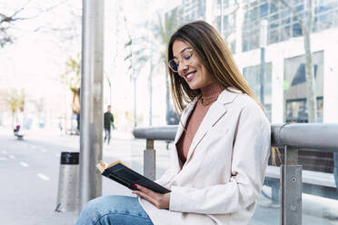 Happy woman reading book sitting at tram station - PNAF03718