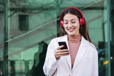 Lächelnde Frau mit Kopfhörern und Smartphone vor einer grünen Glaswand stehend - PNAF03728
