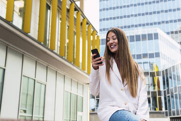 Happy young woman using smart phone sitting on seat in front of building - PNAF03733
