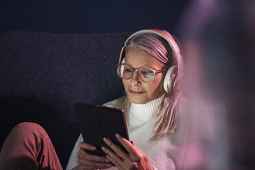 Senior woman with headphones using tablet PC sitting on chair - JOSEF08609