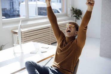 Happy businessman with arms raised shouting sitting at desk in office - JOSEF08690