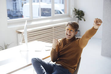 Happy businessman gesturing fist sitting at desk in office - JOSEF08691