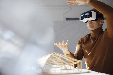 Architect wearing virtual reality simulator gesturing sitting with leaf shaped model at desk in office - JOSEF08693