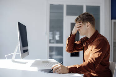 Businessman covering eyes with hands sitting at desk in office - JOSEF08706