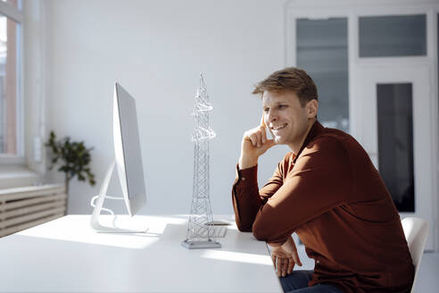 Glücklicher Ingenieur, der ein Modell eines Strommastes am Schreibtisch im Büro betrachtet - JOSEF08713