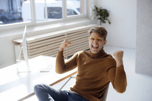 Glücklicher Geschäftsmann mit Brille und gestikulierender Faust, der am Schreibtisch im Büro sitzt - JOSEF08736