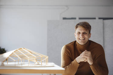 Glücklicher Architekt mit verschränkten Händen, der neben einem blattförmigen Holzmodell im Büro sitzt - JOSEF08739