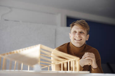 Lächelnder Architekt mit verschränkten Händen, der neben einem blattförmigen Holzmodell im Büro sitzt - JOSEF08740
