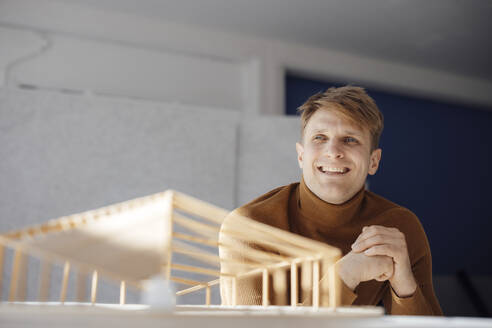 Lächelnder Architekt mit verschränkten Händen, der neben einem blattförmigen Holzmodell im Büro sitzt - JOSEF08740
