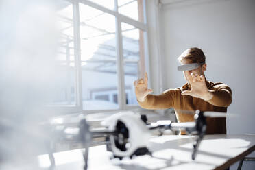 Businessman wearing virtual reality simulator making finger frame sitting with drone at desk - JOSEF08755