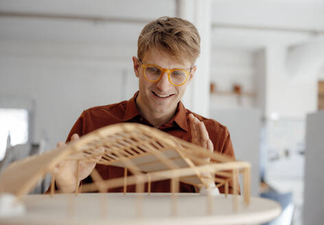 Lächelnder Architekt mit Brille, der ein blattförmiges Modell im Büro betrachtet - JOSEF08777