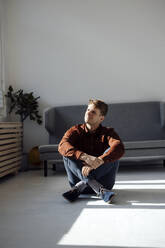 Smiling man sitting on floor in front of sofa in living room at home - JOSEF08785