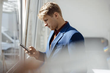 Geschäftsmann mit Smartphone am Fenster im Büro stehend - JOSEF08796