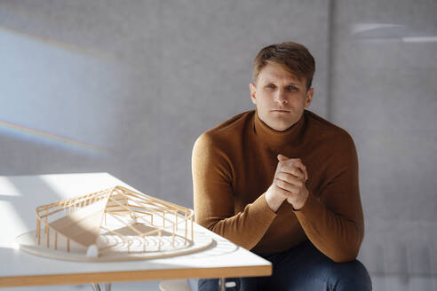 Architekt mit verschränkten Händen, der an einem blattförmigen Holzmodell im Büro sitzt - JOSEF08805