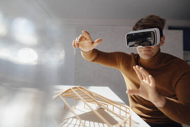 Smiling architect wearing virtual reality simulator gesturing above leaf shaped model at desk in office - JOSEF08807