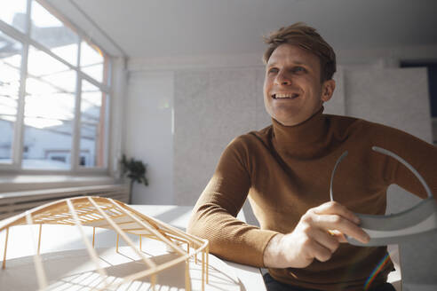 Lächelnder Architekt, der einen Virtual-Reality-Simulator hält und neben einem blattförmigen Modell am Schreibtisch im Büro sitzt - JOSEF08809
