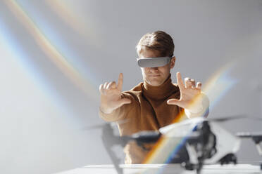 Businessman wearing virtual reality simulator making finger frame towards drone in office - JOSEF08815