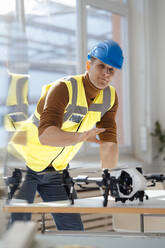 Engineer wearing reflective clothing gesturing with drone at desk in office - JOSEF08819