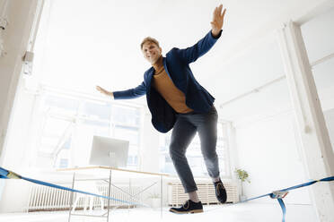 Happy businessman balancing on tightrope in office - JOSEF08820