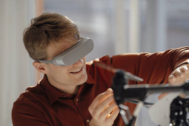 Smiling businessman wearing virtual reality simulator gesturing towards drone in office - JOSEF08823
