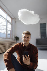 Lächelnder Geschäftsmann, der ein Smartphone hält, das unter einem schwebenden Wolkennetzwerk im Büro sitzt - JOSEF08852