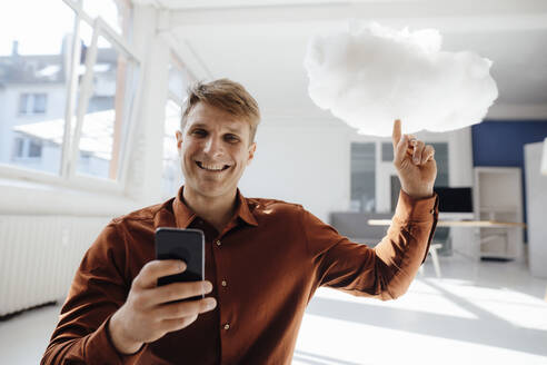 Glücklicher Geschäftsmann, der ein Mobiltelefon hält und auf ein schwebendes Cloud-Netzwerk im Büro zeigt - JOSEF08854