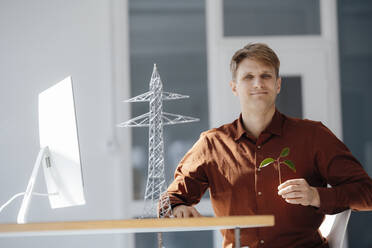 Lächelnder Ingenieur, der ein Blatt in der Hand hält, sitzt neben einem Modell eines Strommastes am Schreibtisch im Büro - JOSEF08865