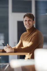 Happy businessman holding mobile phone sitting at desk in office - JOSEF08869