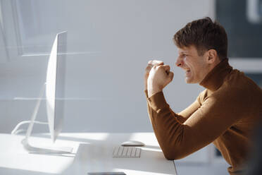 Glücklicher Geschäftsmann mit gestikulierender Faust, der auf einen Desktop-PC im Büro schaut - JOSEF08870