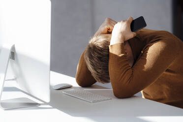 Geschäftsmann hält Handy sitzend mit Händen hinter dem Kopf am Schreibtisch im Büro - JOSEF08873