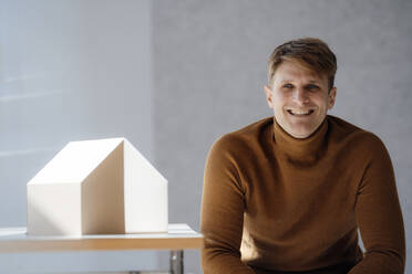 Happy architect sitting by model house in office - JOSEF08879