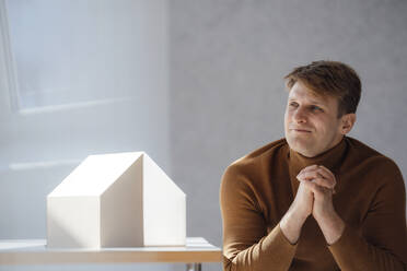 Smiling architect with hands clasped sitting by model house in office - JOSEF08880