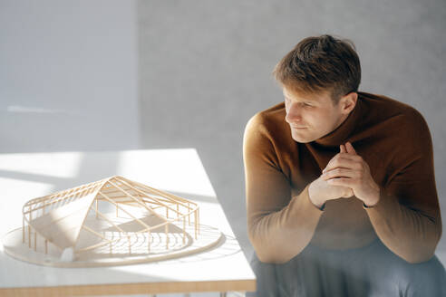 Nachdenklicher Architekt, der ein blattförmiges Modell betrachtet, das am Schreibtisch im Büro sitzt - JOSEF08882