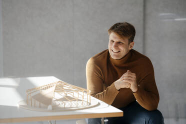 Glücklicher Architekt sitzt mit blattförmigen Modell am Schreibtisch im Büro - JOSEF08883