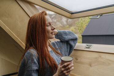 Rothaarige Frau hält Kaffeetasse und schaut durch ein Fenster - OGF01158