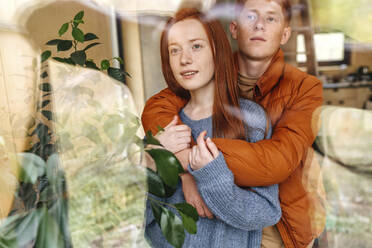 Boy embracing girlfriend from behind seen through glass - OGF01174