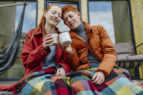 Lächelndes Paar, das auf der Terrasse des Hauses sitzt und eine Kaffeetasse anstößt - OGF01185