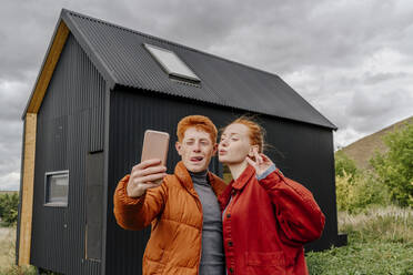 Paar nimmt Selfie durch Smartphone vor winzigem Haus stehend - OGF01194