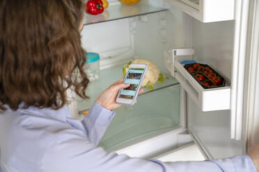 Frau beim Online-Einkauf von Lebensmitteln über ihr Smartphone am Kühlschrank - VPIF05827