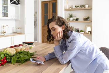 Frau benutzt Mobiltelefon bei Gemüse am Esstisch in der Küche - VPIF05842
