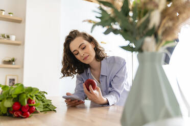 Junge Frau überprüft die Kalorien eines Apfels auf dem Handy in der Küche - VPIF05844
