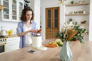 Young woman peeling carrot at dining table in kitchen - VPIF05847
