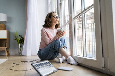 Junge Geschäftsfrau mit Kaffeetasse, die durch das Fenster ihres Hauses schaut - VPIF05879