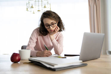 Studentin schreibt in Buch an Laptop am Tisch - VPIF05882