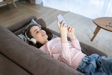 Young woman using mobile phone lying on sofa in living room - VPIF05905