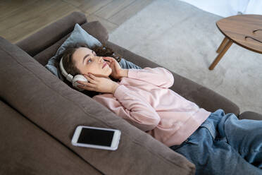 Junge Frau hört Musik über Kopfhörer auf dem Sofa zu Hause - VPIF05906