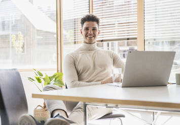 Smiling young businessman with laptop at desk in office - UUF25741