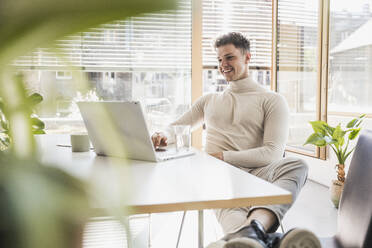 Smiling young businessman using laptop at desk in office - UUF25745