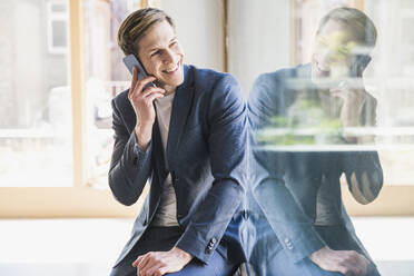 Lächelnder Geschäftsmann am Telefon am Fenster im Büro - UUF25773