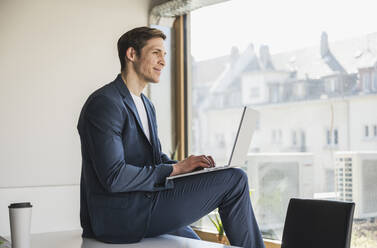 Businessman using laptop at the window in office - UUF25778